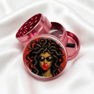 May include: Pink metal herb grinder with a Medusa graphic on the lid. The graphic features a woman with sunglasses blowing a bubblegum bubble. The grinder is disassembled, showing the internal components.