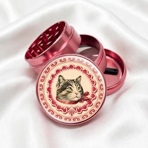 Peut inclure: Un moulin à herbes rose, démonté. La partie supérieure révèle les dents de broyage. La base présente un portrait de chat avec un ruban rouge. Conçu pour les herbes et les épices.