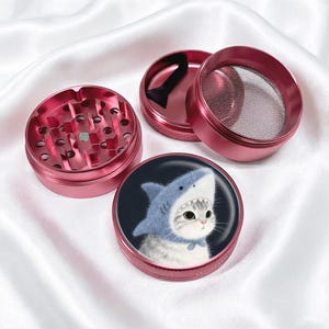 May include: A pink, four-piece herb grinder. The top piece has holes for grinding, and the bottom piece features a cat wearing a shark hat. The grinder is made of metal and has a mesh screen.
