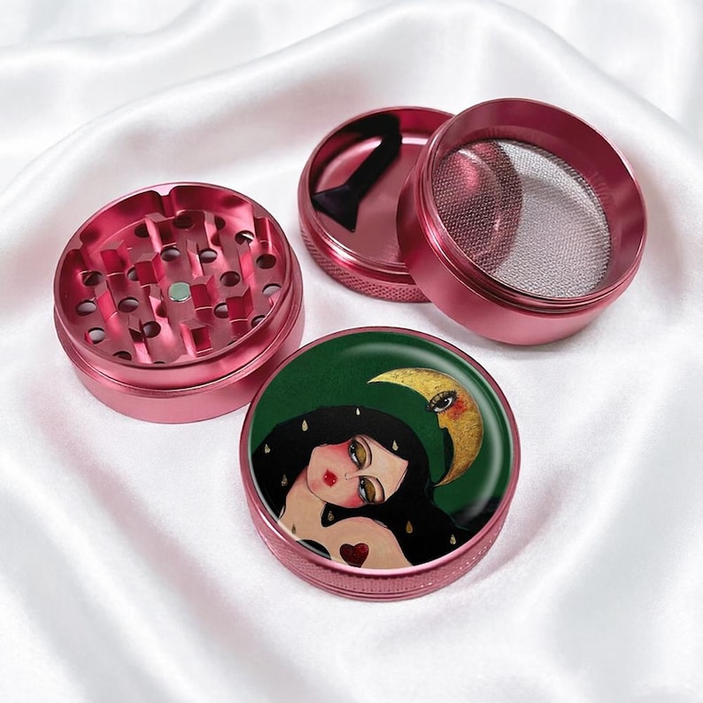 May include: A disassembled pink metal herb grinder with a decorative top featuring a woman with long black hair, a red heart, and a crescent moon on a green background. The grinder is on a white satin surface.