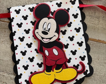Op de afbeelding: Een decoratieve banner-hanger met een gelaagde papieren uitsparing van Mickey Mouse in een rode broek en gele schoenen. De achtergrond is wit met een herhalend patroon van zwarte Mickey Mouse-hoofden en kleine veelkleurige stippen, omlijst door een zwarte geschulpte rand.