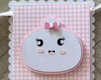 May include: A rectangular paper banner with a scalloped white border and a pink and white gingham pattern. Centered on the banner is a white, rounded character with a smiling face, black eyes, pink cheeks, and a small pink bow with a pearl accent.