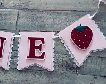 May include: A pink banner with the word "ONE" and a strawberry illustration. The letters are burgundy with white outlines, and the strawberry is red with a green stem. The banner has a scalloped edge.