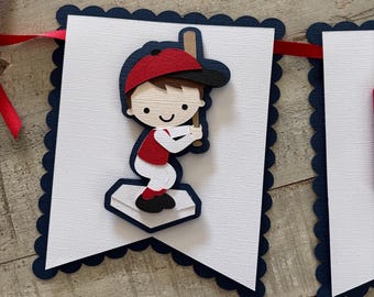 Op de afbeelding: Een decoratieve banner met een gelaagde papieren uitsparing van een honkbalspeler met een rode pet, rood shirt en witte broek, die een beige knuppel vasthoudt terwijl deze op een witte honkplaat staat, gemonteerd op een witte kaart met marineblauwe rand.