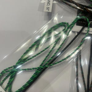 May include: Close-up of several archery bowstrings in clear plastic packaging. The bowstrings are various colors, including green, black, and brown. The text on the packaging reads "97, 15 STRAND, 50".