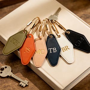 May include: A collection of six diamond-shaped hotel-style key tags in olive green, orange, light pink, navy blue, white, and black. Each tag features gold-embossed initials and is attached to a gold-toned metal wire ring. A silver key is included.