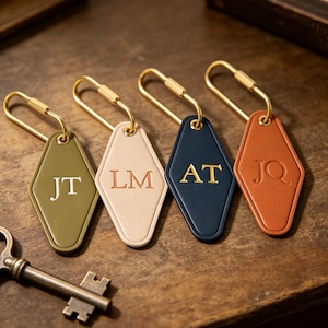Può includere: Quattro portachiavi in pelle a forma di diamante nei colori verde oliva, beige, blu navy e terracotta, dotati di finiture in metallo dorato e monogrammi a due lettere in rilievo: JT, LM, AT e JQ. Una chiave antica in metallo è vicina.
