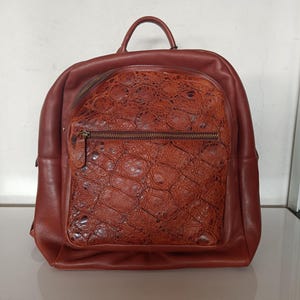 Può includere: Uno zaino in pelle marrone con una tasca frontale strutturata e una maniglia superiore. Lo zaino ha una tasca frontale con cerniera e uno scomparto principale. La pelle ha una leggera lucentezza e il design è classico e funzionale.