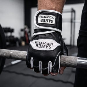 May include: A black and white fingerless weightlifting glove featuring a wide wrist strap with a hook-and-loop closure. The glove displays the text SAHER INDUSTRIES on both the wrist strap and the white mesh panel on the back of the hand.