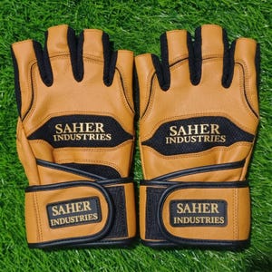 May include: A pair of fingerless gloves in tan leather and black mesh fabric. The gloves feature black trim and a black wrist strap with a rectangular patch displaying the text SAHER INDUSTRIES in gold lettering. The same text appears on the back.