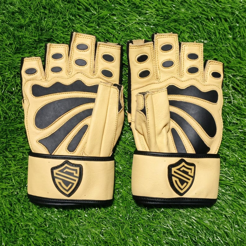 May include: A pair of fingerless gloves in beige and black. The gloves feature black rubberized padding on the fingers and palm area, with a beige wrist strap displaying a black shield-shaped logo with a stylized letter S in the center.