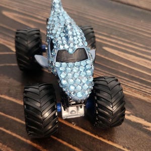 May include: A custom monster truck toy with a shark-like design. The light blue body is covered in sparkling blue rhinestones. It has large black tyres and a blue chassis, set against a wooden surface.