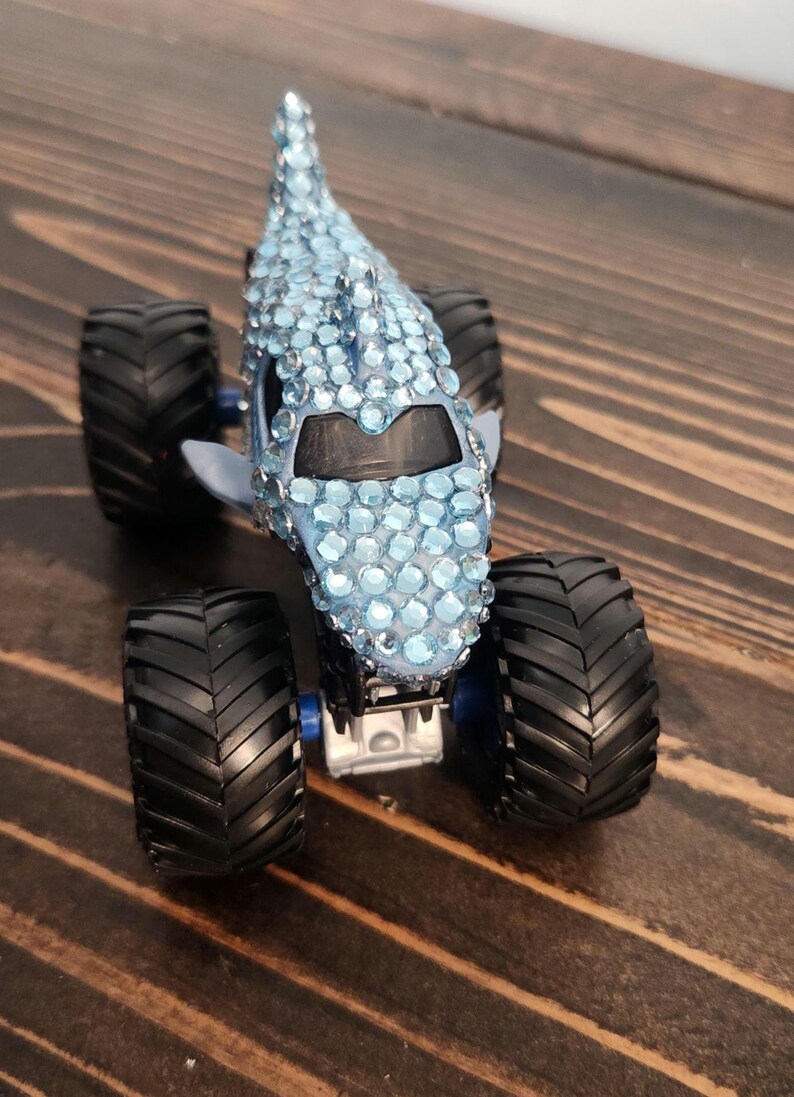 May include: A custom monster truck toy with a shark-like design. The light blue body is covered in sparkling blue rhinestones. It has large black tyres and a blue chassis, set against a wooden surface.