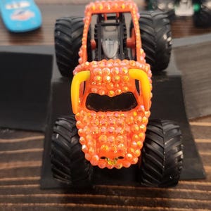 May include: Orange monster truck toy with black tires and yellow accents. The truck is covered in sparkling embellishments. Other toy vehicles are visible in the background.
