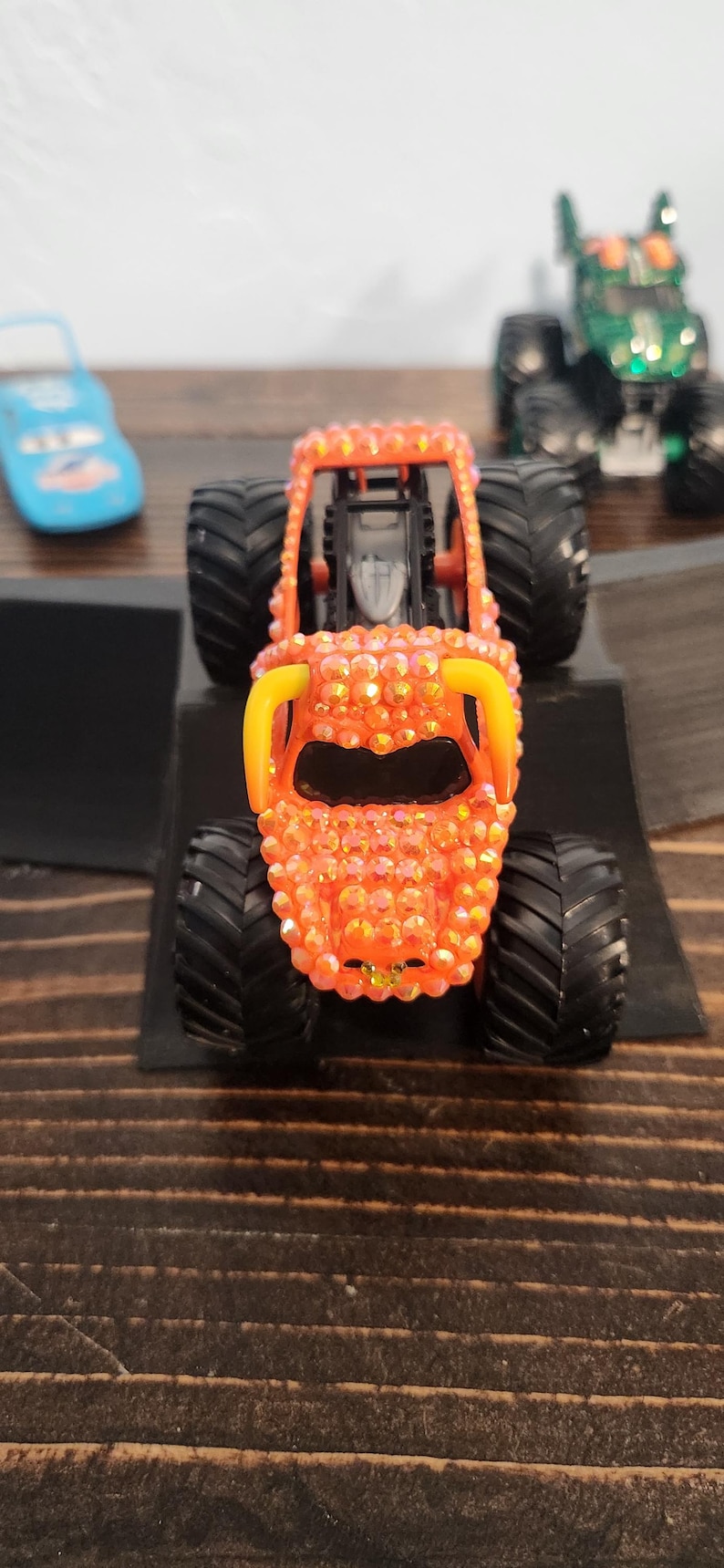 May include: Orange monster truck toy with black tires and yellow accents. The truck is covered in sparkling embellishments. Other toy vehicles are visible in the background.
