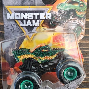 May include: A Monster Jam Dragon truck toy in a clear plastic package. The truck is green, orange, and black with large black wheels and green hubs. The packaging features the Monster Jam logo and the words "Legacy Trucks".