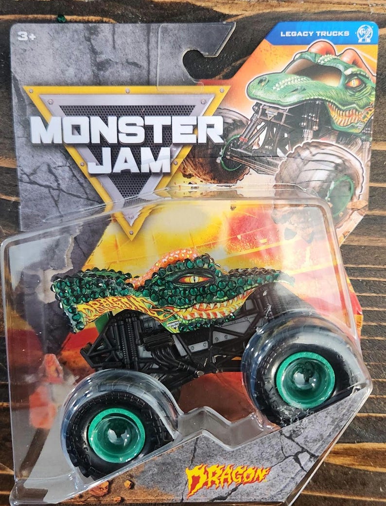 May include: A Monster Jam Dragon truck toy in a clear plastic package. The truck is green, orange, and black with large black wheels and green hubs. The packaging features the Monster Jam logo and the words "Legacy Trucks".