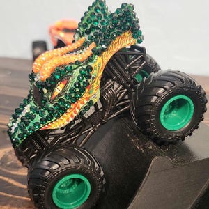 May include: A toy monster truck with a dragon-themed design. The truck features a green and orange color scheme, with green gemstones covering the dragon's head. The wheels are black with green rims. The truck is positioned on a black ramp.
