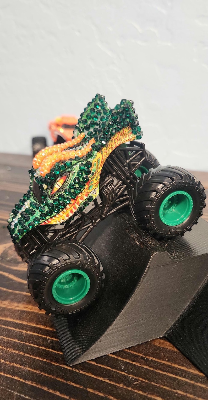 May include: A toy monster truck with a dragon-themed design. The truck features a green and orange color scheme, with green gemstones covering the dragon's head. The wheels are black with green rims. The truck is positioned on a black ramp.