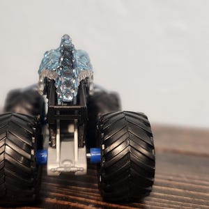 May include: A black monster truck toy with large, textured tyres and a silver and blue decorative top. The toy is set against a neutral background, highlighting its details.