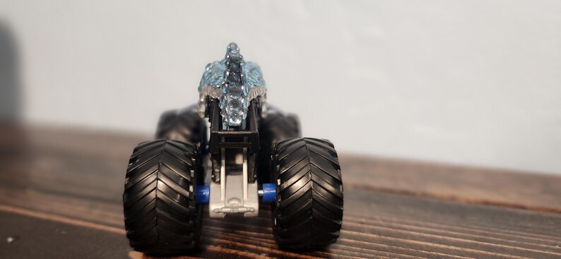 May include: A black monster truck toy with large, textured tyres and a silver and blue decorative top. The toy is set against a neutral background, highlighting its details.
