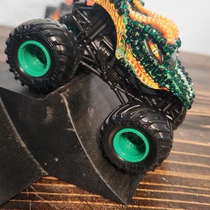 May include: A toy monster truck with a dragon design. The truck has large black tires with green rims. The dragon is green, orange, and yellow with crystal-like accents. The truck is on a black ramp.