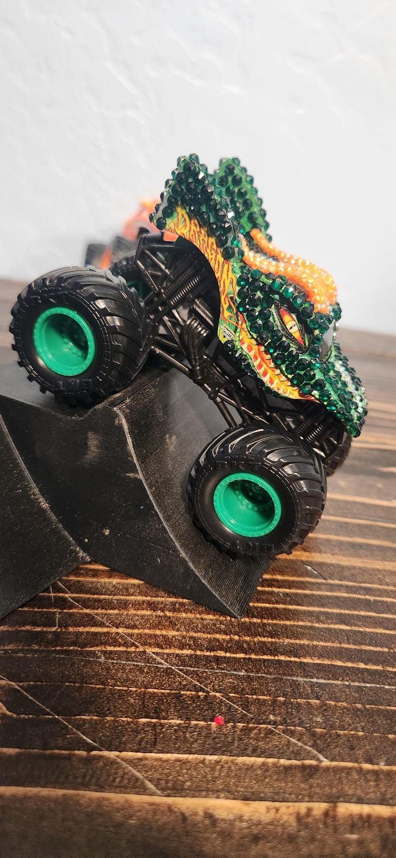 May include: A toy monster truck with a dragon design. The truck has large black tires with green rims. The dragon is green, orange, and yellow with crystal-like accents. The truck is on a black ramp.