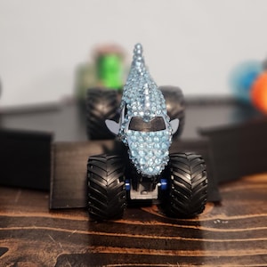 May include: A selection of toy monster trucks, with a central truck covered in blue rhinestones. Other trucks in the background are orange, green, and brown. The trucks are on black ramps, set against a wooden surface.