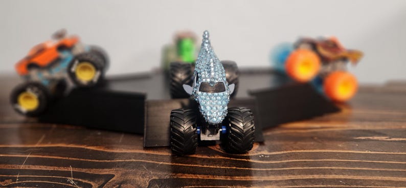 May include: A selection of toy monster trucks, with a central truck covered in blue rhinestones. Other trucks in the background are orange, green, and brown. The trucks are on black ramps, set against a wooden surface.
