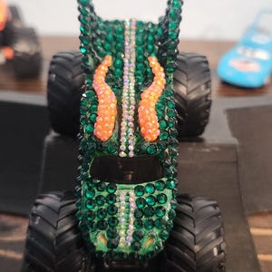 May include: A green monster truck toy covered in sparkling green and iridescent rhinestones. The truck features orange accents and large black tires. A blue toy car is visible in the background.