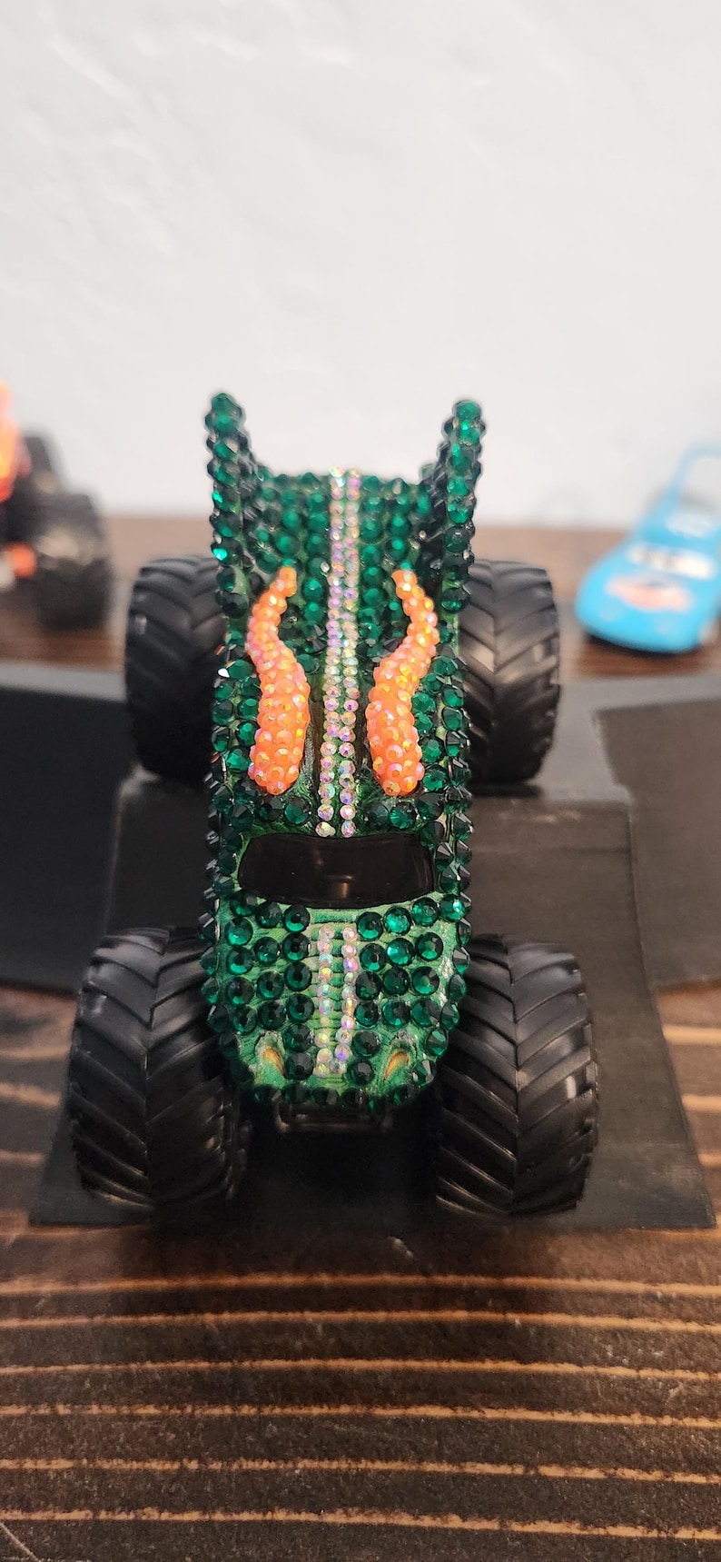 May include: A green monster truck toy covered in sparkling green and iridescent rhinestones. The truck features orange accents and large black tires. A blue toy car is visible in the background.
