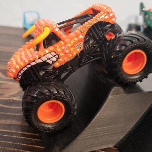 May include: Orange monster truck toy with black wheels and orange accents. The truck has a bull-like design with the words "El Toro Loco" on the side. The wheels are large and the truck is on a ramp.