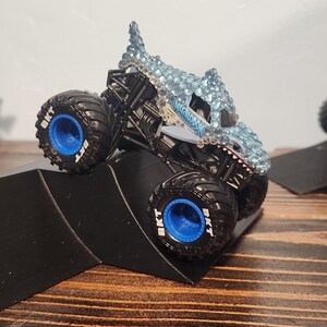 May include: Three toy monster trucks with large tyres are positioned on black ramps. The trucks have different designs, including a blue and silver truck, a brown and orange truck, and a red truck with orange wings. The trucks are on a wooden surface.