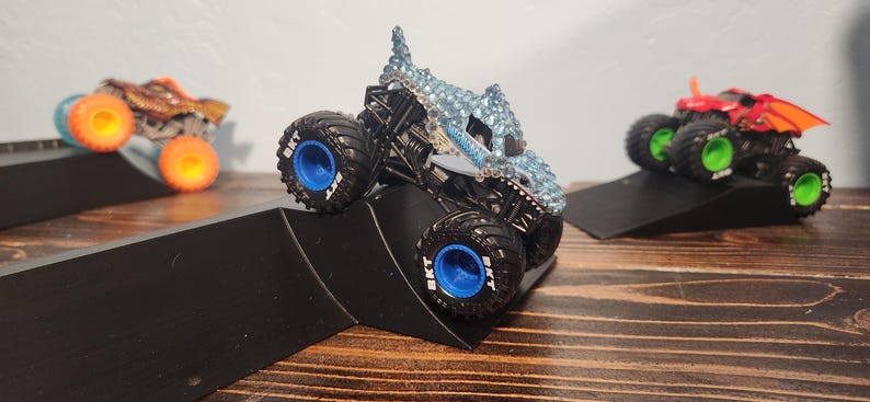 May include: Three toy monster trucks with large tyres are positioned on black ramps. The trucks have different designs, including a blue and silver truck, a brown and orange truck, and a red truck with orange wings. The trucks are on a wooden surface.