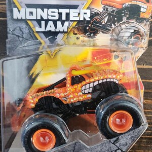 May include: Orange and yellow El Toro Loco monster truck toy in a clear plastic package. The truck has large black tires with orange rims and a detailed design. The packaging includes the "Monster Jam" logo and "Legacy Trucks" text.