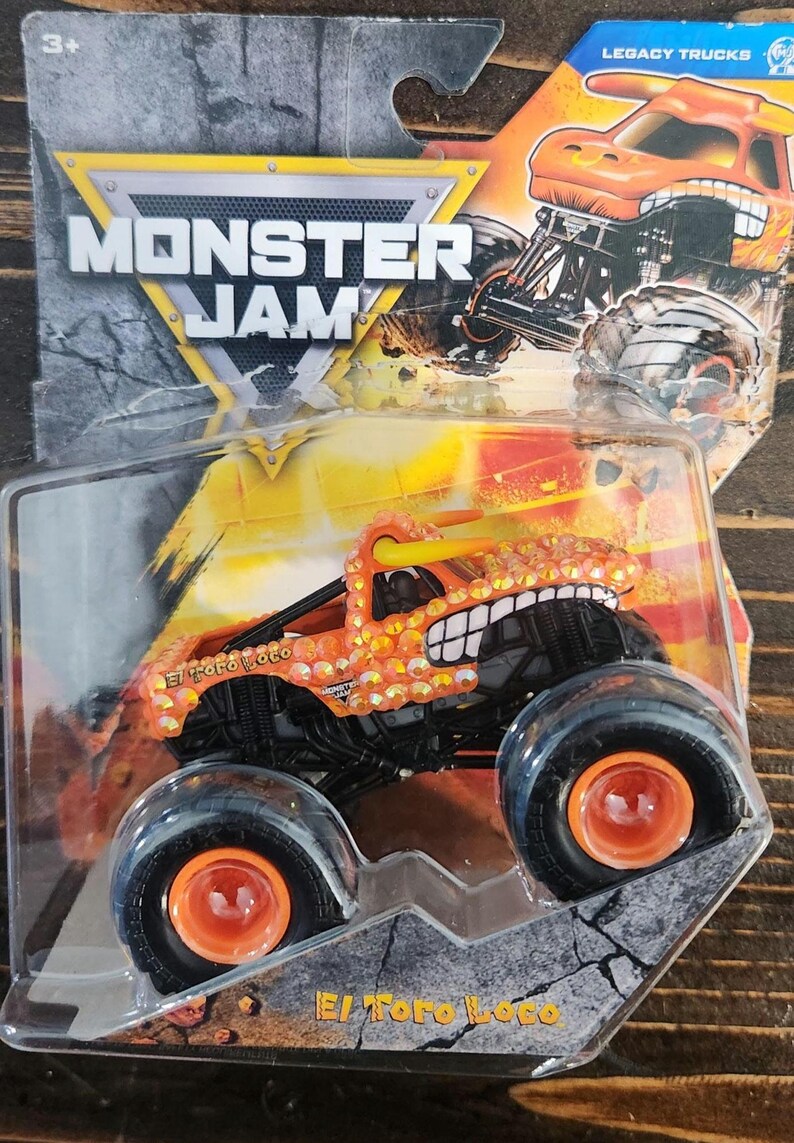 May include: Orange and yellow El Toro Loco monster truck toy in a clear plastic package. The truck has large black tires with orange rims and a detailed design. The packaging includes the "Monster Jam" logo and "Legacy Trucks" text.