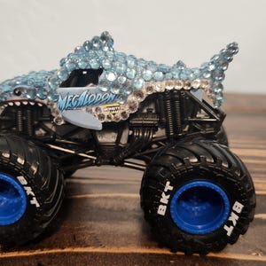 May include: A toy monster truck with a shark design. The truck has a light blue shark head and body covered in rhinestones, black tyres with blue rims, and the word "MEGALODON" on the side.