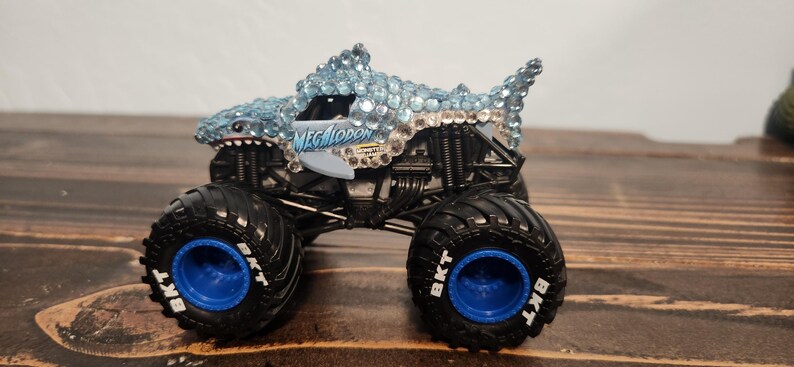 May include: A toy monster truck with a shark design. The truck has a light blue shark head and body covered in rhinestones, black tyres with blue rims, and the word "MEGALODON" on the side.
