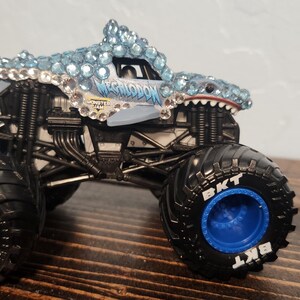 May include: A toy monster truck with a shark-themed design. The truck has a shark head with blue and clear gem-like accents. The tyres are black with blue rims and the text "BKT".