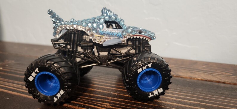 May include: A toy monster truck with a shark-themed design. The truck has a shark head with blue and clear gem-like accents. The tyres are black with blue rims and the text "BKT".