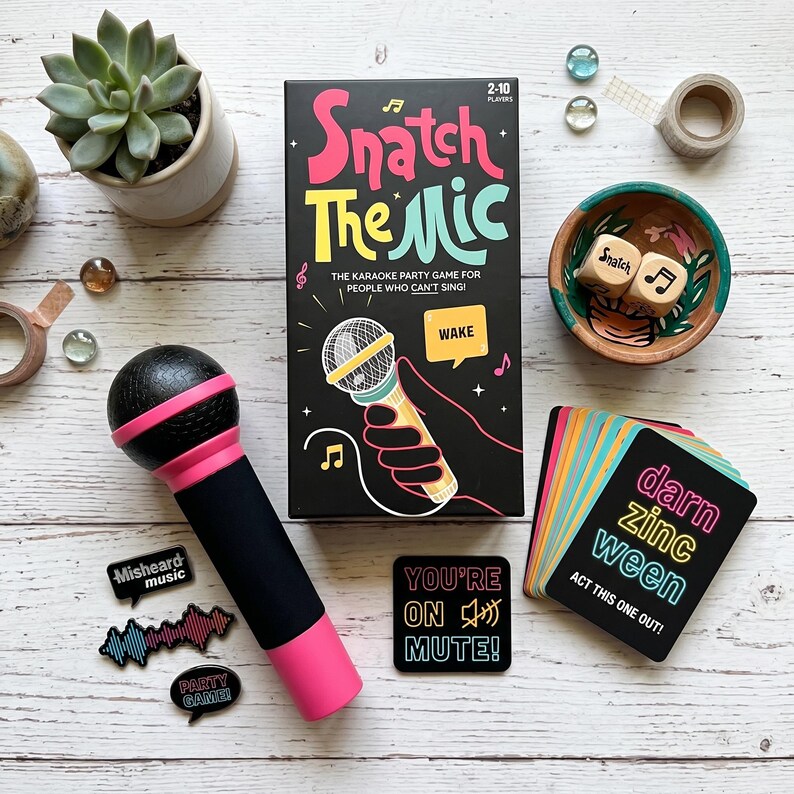 May include: A karaoke party game set, "Snatch The Mic," includes a black box with colorful text, a toy microphone, cards, and dice. The game box features a microphone graphic and the text "The Karaoke Party Game for People Who Can't Sing!"