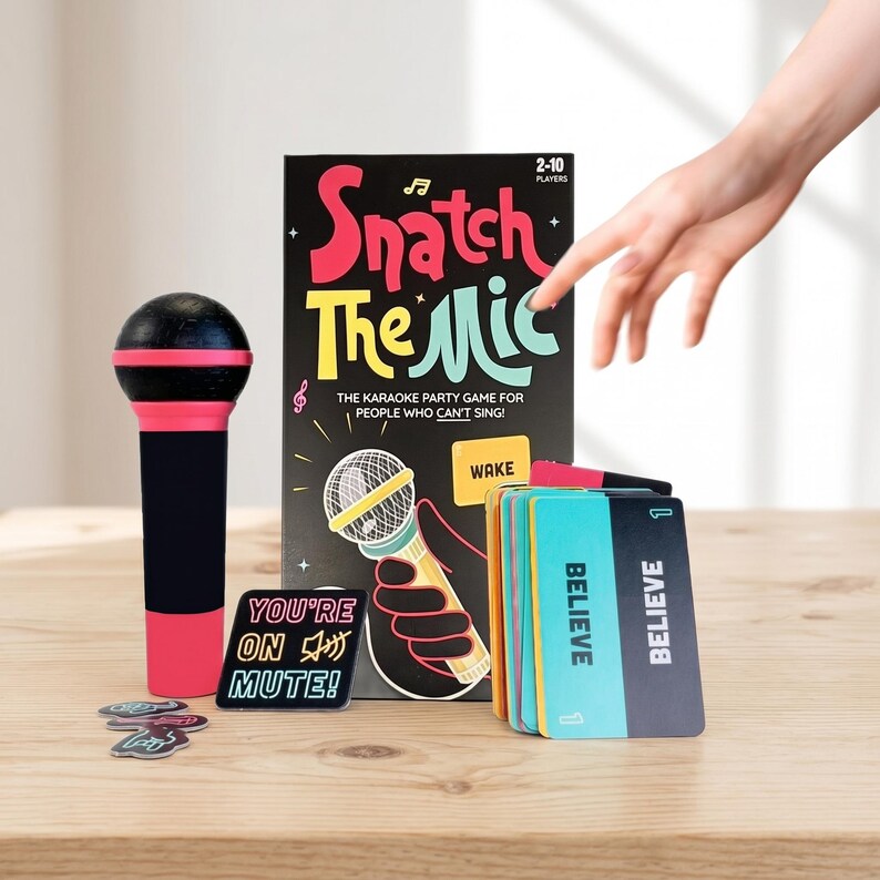May include: A karaoke party game called "Snatch The Mic" with a black and pink microphone prop, cards, and game pieces. The game box features a microphone illustration and the text "The Karaoke Party Game For People Who Can't Sing!"