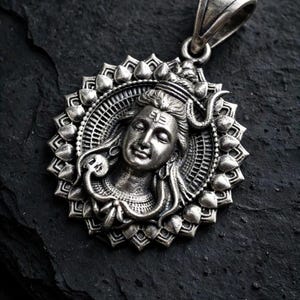 May include: A circular silver pendant featuring a detailed relief of a deity with a third eye on the forehead, surrounded by a stylised lotus petal border and a snake coiled around the neck, topped with a sturdy triangular bail for hanging.