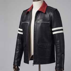 Könnte beinhalten: Eine schwarze Lederjacke mit einem kontrastierenden roten Kragen, zwei weißen horizontalen Streifen an jedem Ärmel, silberfarbenen Reißverschlusstaschen auf der Brust und zwei unteren Pattentaschen. Die Jacke hat einen durchgehenden Frontreißverschluss.