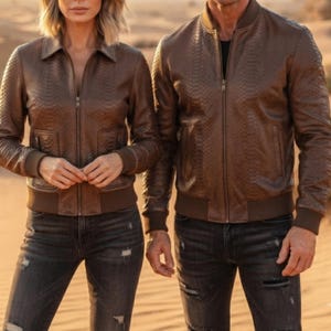 May include: Two brown leather bomber jackets featuring a textured snakeskin pattern, full-length front zippers, ribbed cuffs, and ribbed waistbands. Both jackets are paired with dark gray distressed denim jeans with visible rips and frayed details.