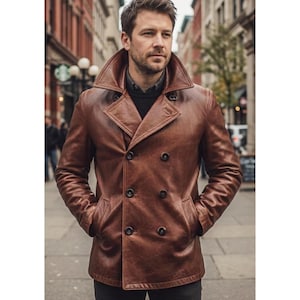 Puede incluir: Un chaquetón de cuero marrón de doble botonadura con un cuello ancho de solapas, mangas largas y botones oscuros. La prenda se lleva sobre un suéter oscuro, mostrando un corte estructurado con costuras visibles y una textura de cuero liso.
