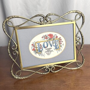 May include: A rectangular brass picture frame with an ornate, swirling metal border featuring floral details. Inside the frame is a light gray mat surrounding an oval print that says LOVE ALWAYS AND FOREVER in blue and black with red roses.