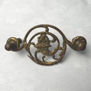 May include: A brass drawer pull featuring a circular frame with an openwork design. The center depicts a cornucopia overflowing with fruit. The handle has two mounting points on either side, each finished with a rounded, knob-like end piece.