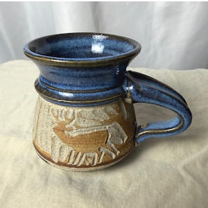 May include: A ceramic mug featuring a flared rim with a glossy blue glaze. The lower body has a textured, unglazed tan surface with a carved relief of a deer. The handle is finished in a matching glossy blue glaze, extending from the rim to the base.