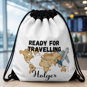 Può includere: Uno zaino a sacco bianco con una stampa nera che riporta la scritta READY FOR TRAVELLING e il nome Holger. Il design include una mappa del mondo color marrone chiaro, l'illustrazione di un aereo blu e rotte di volo tratteggiate in nero.